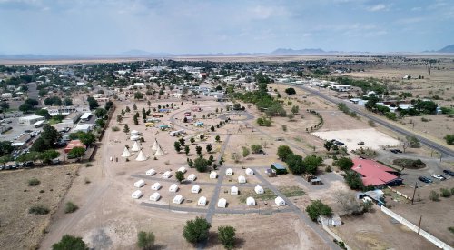 Aerial view of a small town with tents and tipis, surrounded by arid land and distant mountains under a cloudy sky.