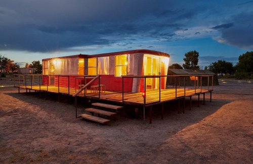 A red trailer home with warm lights, set on a wooden deck, surrounded by a sandy area at dusk.