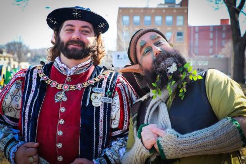 Two men in colorful costumes, one in a regal outfit and the other as a whimsical elf, smiling outdoors.