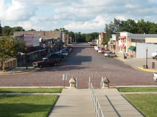 A quiet, tree-lined street with brick pavement, featuring shops and parked cars under a partly cloudy sky.