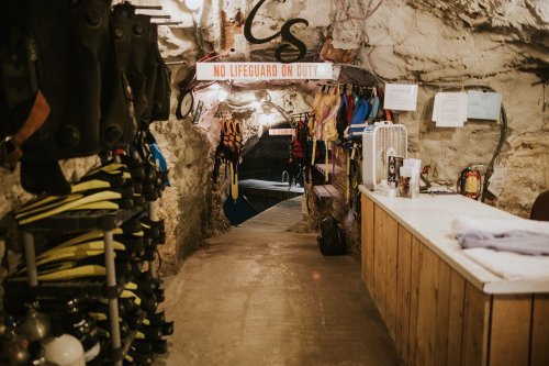 Interior of a cave with diving gear on the left, a sign reading "No Lifeguard on Duty," and a pool area ahead.