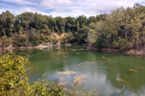 A serene green pond surrounded by trees and rocky terrain under a partly cloudy sky.