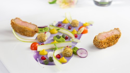 A gourmet dish featuring two pieces of breaded meat on a vibrant purple sauce, garnished with colorful vegetables and sauces.