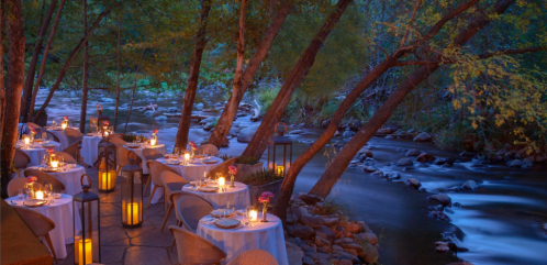 Romantic outdoor dining setup by a river, with tables, candles, and soft lighting amidst trees at dusk.