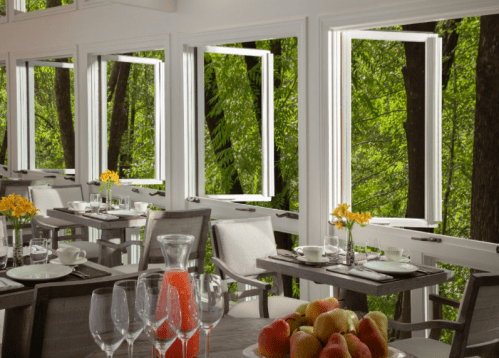 A bright restaurant with open windows overlooking lush greenery, featuring tables set with dishes and a fruit bowl.