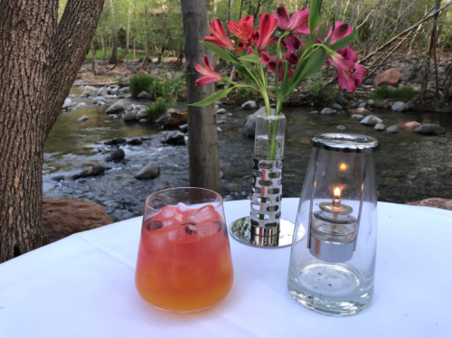 A refreshing cocktail sits on a table beside a candle and a vase of flowers, with a serene river in the background.