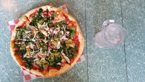 A pizza topped with spinach, onions, and peppers, next to a glass of water on a green textured table.