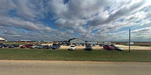 A wide view of a parking lot filled with cars, with a large building and a cloudy sky in the background.