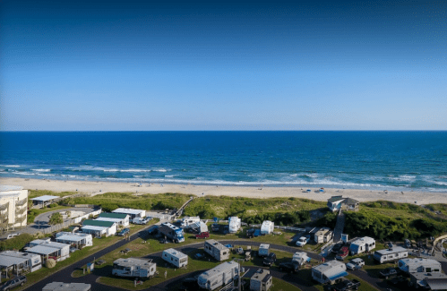 Aerial view of a beach with waves, surrounded by a campground and RVs, under a clear blue sky.