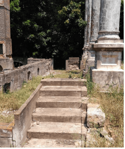 Stone steps lead up to weathered columns amidst overgrown grass and trees in a historic ruin.