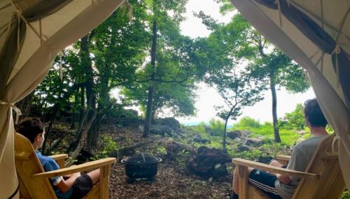 Two people sit in wooden chairs inside a tent, facing a scenic view of trees and a distant body of water.