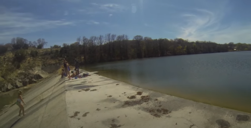 A serene lakeside scene with people enjoying the sun on a concrete shore, surrounded by trees and clear blue skies.
