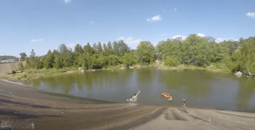 A person slides into a pond while another person prepares to use a float nearby, surrounded by trees and blue sky.