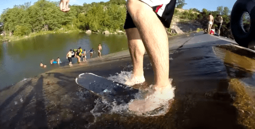 A person skateboarding down a wet ramp into a body of water, with others enjoying the area in the background.