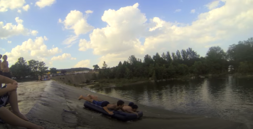 Two boys slide down a slope on a raft into a river, with spectators sitting nearby under a cloudy sky.