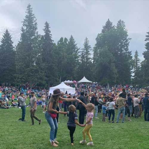 A lively outdoor gathering with people dancing on grass, surrounded by trees and a large crowd in the background.