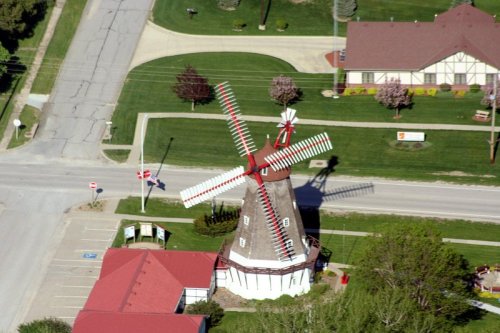 Aerial view of a red and white windmill near a road, surrounded by green grass and a few buildings.