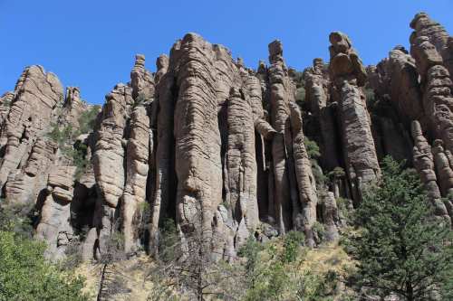 Tall, rugged rock formations rise against a clear blue sky, surrounded by sparse greenery and rocky terrain.