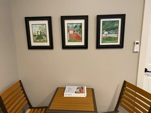 A small table with wooden chairs under three framed landscape paintings on a gray wall. A book rests on the table.