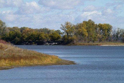 A serene river scene with trees along the shore and a cloudy blue sky reflecting on the water's surface.