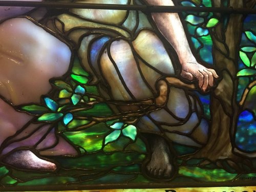 Stained glass artwork featuring a figure seated among vibrant green leaves and colorful backgrounds.