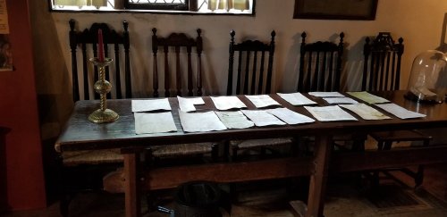 A wooden table with scattered papers and a candle holder, surrounded by tall, dark wooden chairs in a historic room.