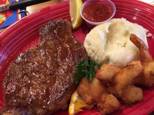A plate featuring a juicy steak, mashed potatoes, fried shrimp, a lemon wedge, and a small cup of dipping sauce.