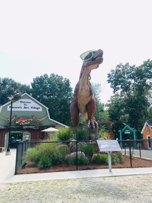 A large dinosaur statue wearing sunglasses stands in front of Nature's Art Village, surrounded by greenery and a fence.
