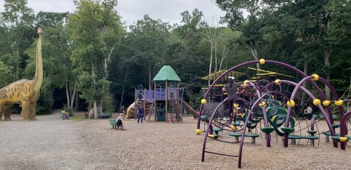 A playground with dinosaur sculptures, climbing structures, and children playing among trees in a natural setting.