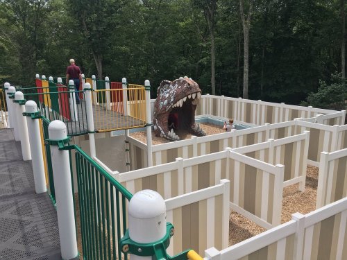 A playground maze featuring a large dinosaur head entrance, surrounded by colorful barriers and trees in the background.