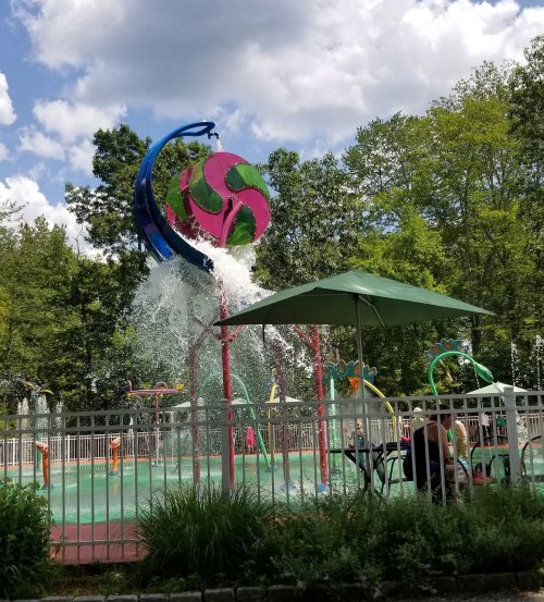 A colorful water play area with a large splash feature, surrounded by greenery and a shaded seating area.