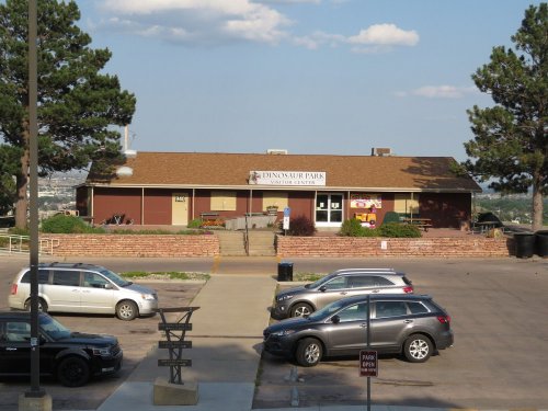 A visitor center at Dinosaur Park, surrounded by trees and parked cars, with a clear blue sky in the background.