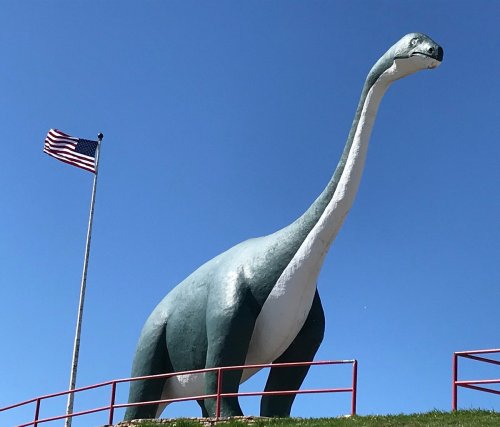 A large dinosaur statue stands against a clear blue sky, with an American flag waving nearby.
