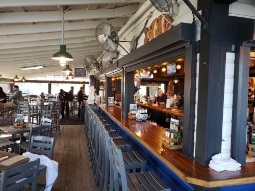 Interior of a casual restaurant with a bar, tables, and patrons, featuring ceiling fans and a wooden floor.