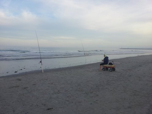 A person sits on a beach with fishing rods set up in the sand, overlooking the ocean under a cloudy sky.