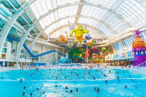 A vibrant indoor water park with large inflatable characters, a wave pool, and numerous visitors enjoying the water.