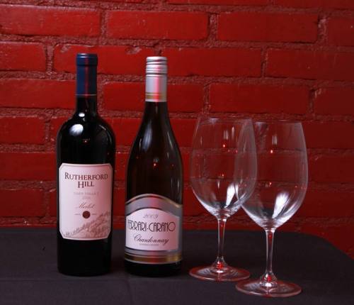 Two wine bottles, a red and a white, beside two empty wine glasses on a black table against a red brick background.