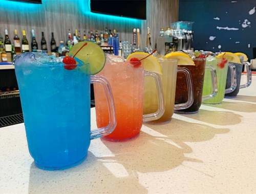 Five colorful pitchers of cocktails lined up on a bar, each garnished with cherries and citrus slices.