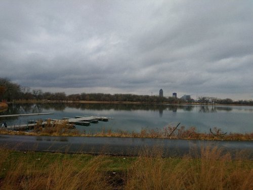 A calm lake reflects a cloudy sky, with a distant city skyline and grassy shoreline in the foreground.