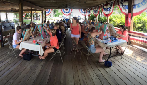A group of women painting on canvases at a covered outdoor space decorated with red, white, and blue bunting.
