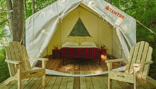 A cozy glamping tent with a bed, surrounded by trees and two wooden chairs on a deck.