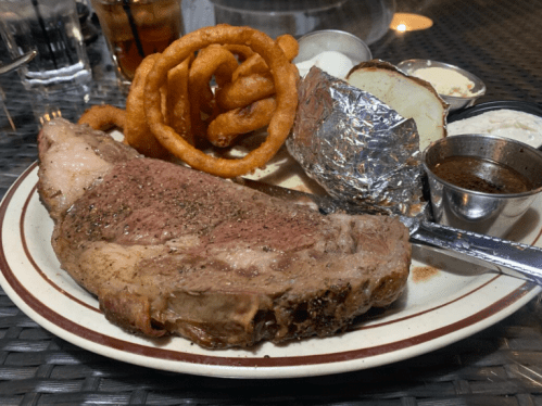 A plate featuring a large prime rib steak, curly fries, a baked potato, and dipping sauces.