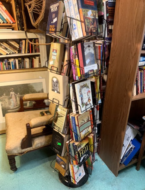 A tall book rack filled with various books, next to a vintage chair in a cozy bookstore setting.
