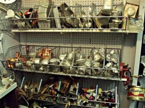 A cluttered display of various metal kitchen tools and utensils organized in wire baskets on a pegboard wall.
