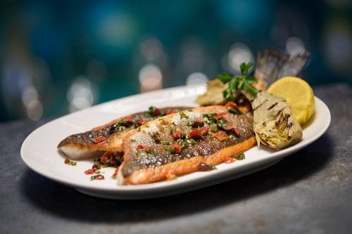 Grilled fish fillet garnished with herbs and lemon, served on a white plate with a side of grilled vegetables.