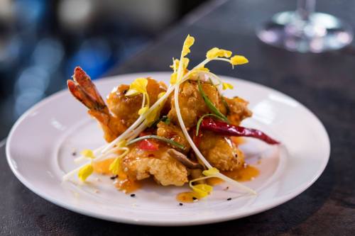 A plate of crispy fried shrimp garnished with herbs, chili peppers, and yellow flowers, drizzled with sauce.