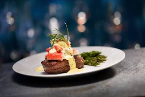 A gourmet steak topped with sauce and garnished, served with asparagus on a white plate against a blurred background.
