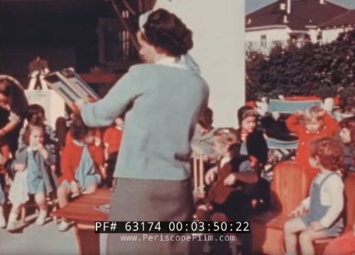A woman reads to a group of young children seated outdoors, with colorful clothing and a sunny setting.