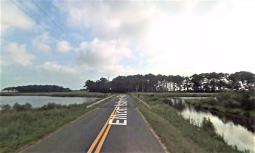 A two-lane road named Elliott Island Road runs alongside a body of water, bordered by trees under a cloudy sky.