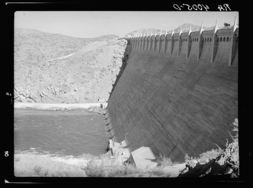 A large concrete dam with a sloped face, surrounded by rocky terrain and a river below, under a clear sky.